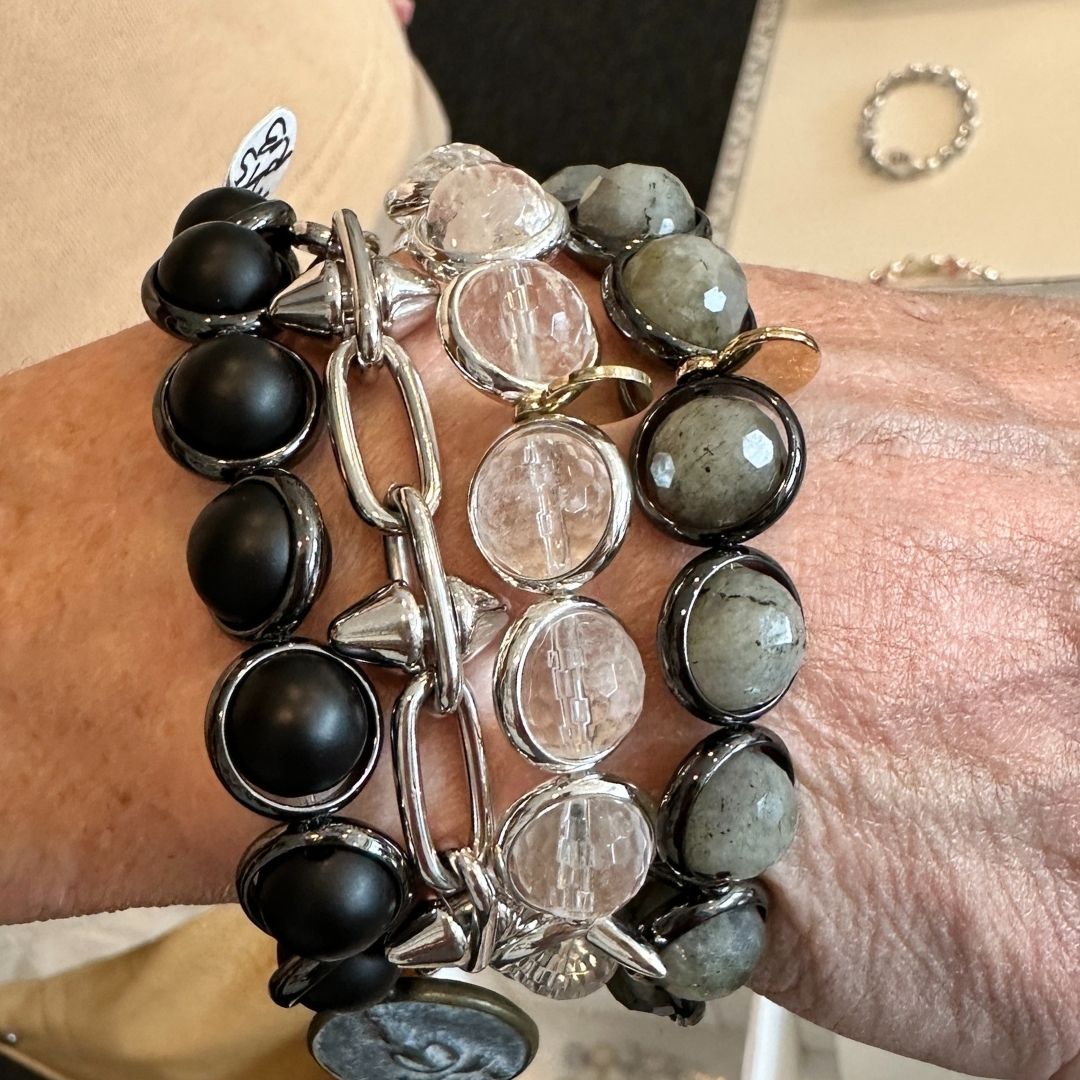 A wrist wearing stacked black and clear gemstone bracelets by Brett Lauren, shown next to a jewelry tray filled with beads and gemstones.