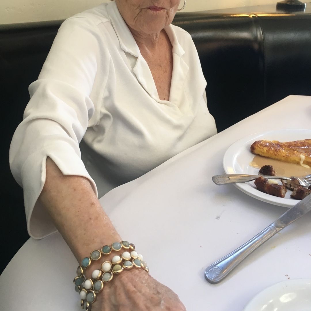 A woman wearing multiple handcrafted gemstone bracelets by Brett Lauren while seated at a table, showcasing elegant jewelry in a casual dining setting.