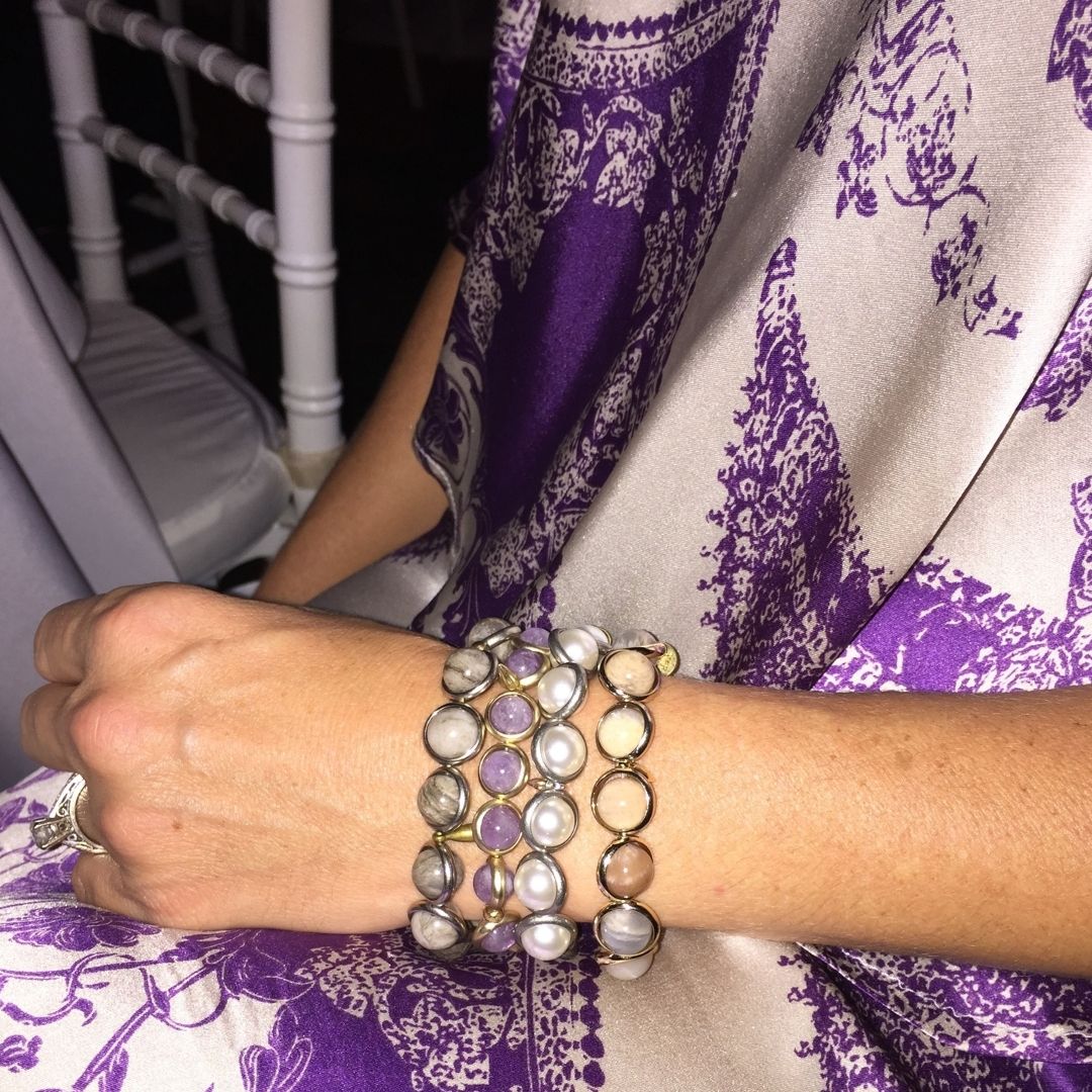 A woman wearing a luxurious collection of Brett Lauren handcrafted gemstone bracelets, styled with a purple floral dress.