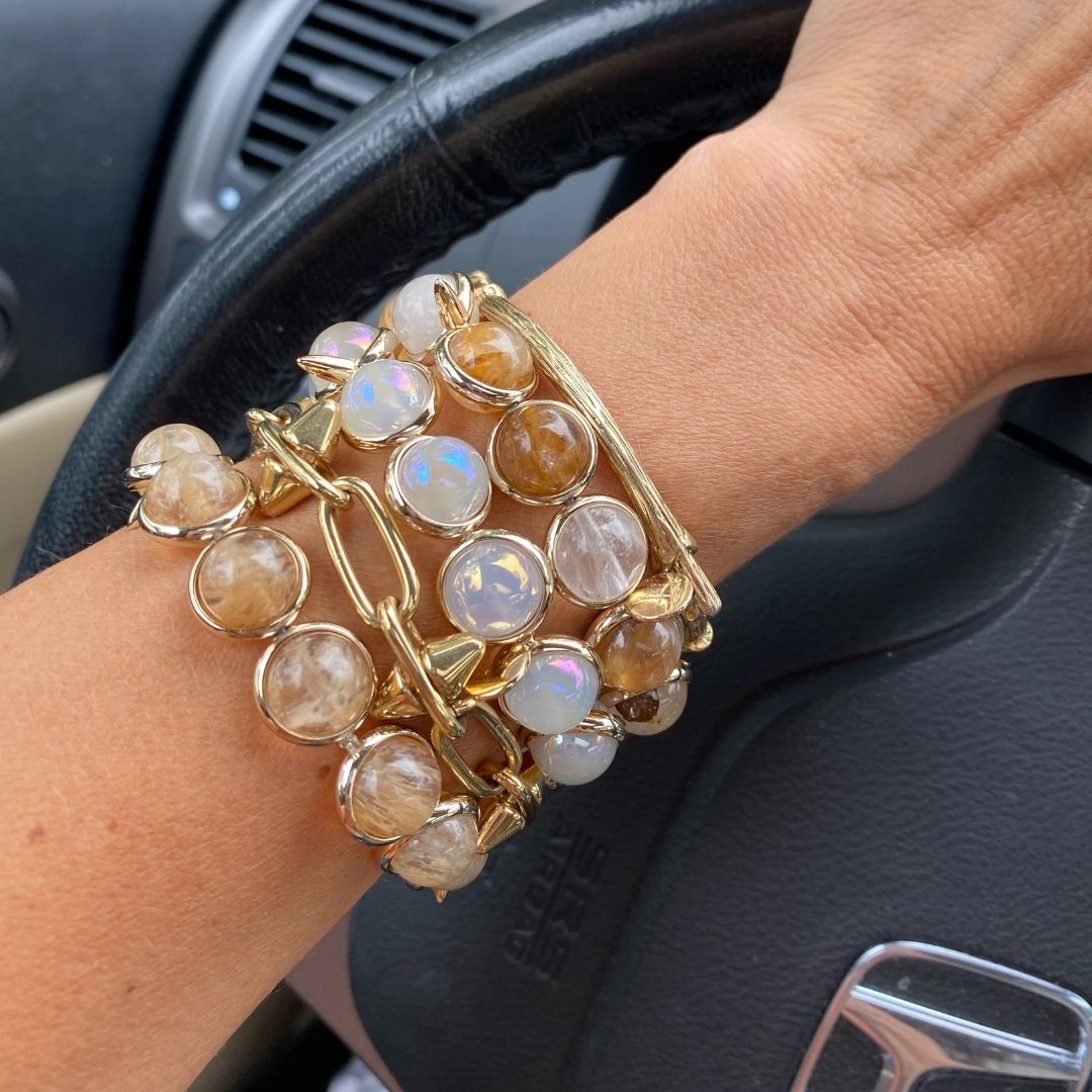 A close-up of a wrist wearing stacked gold and white gemstone bracelets by Brett Lauren while holding a steering wheel, styled in a driving setting.