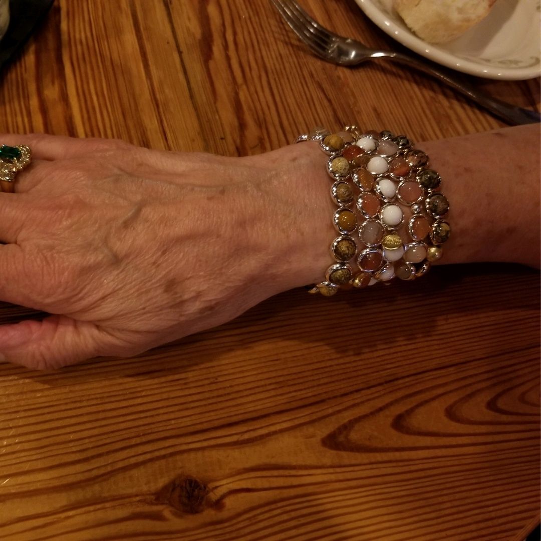 A close-up of a hand wearing stacked gemstone bracelets by Brett Lauren at a restaurant table, with a wooden surface and casual dining setting.