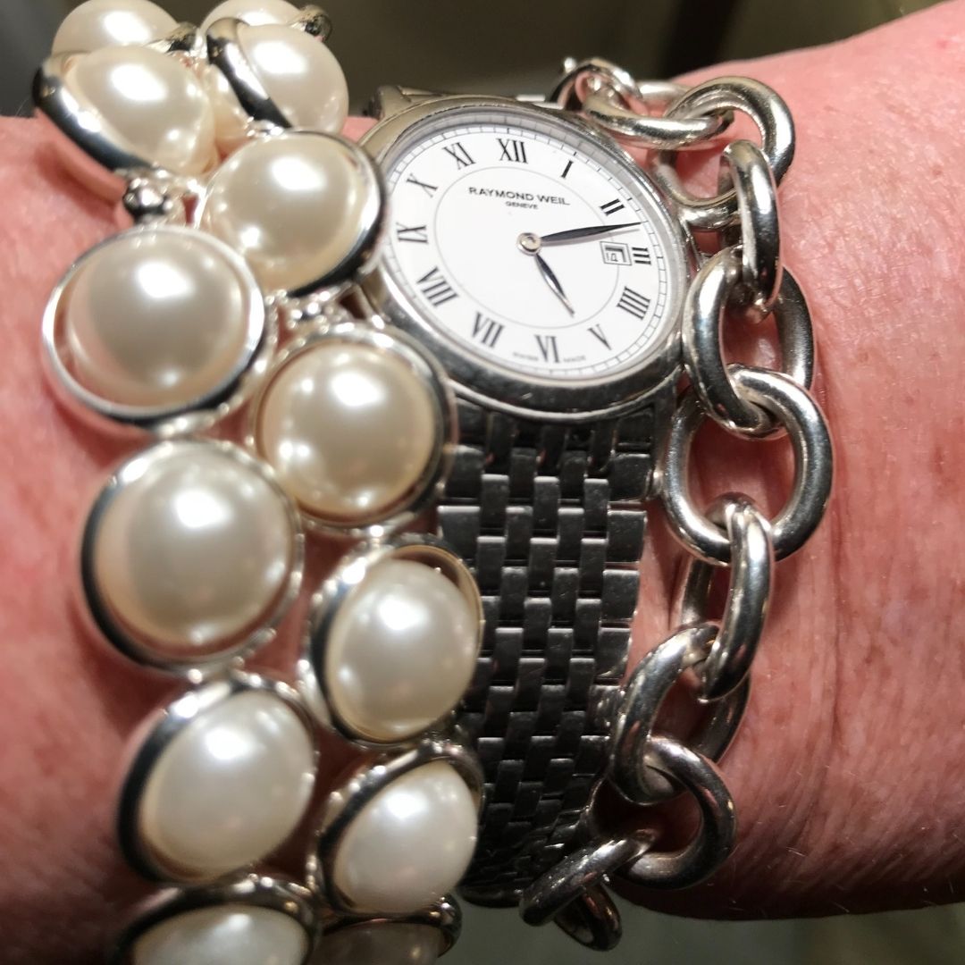 A wrist wearing stacked pearl bracelets by Brett Lauren, paired with a silver watch, showcasing a mix of elegance and casual style.