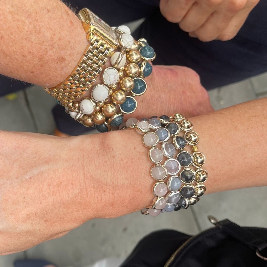 Two wrists adorned with multiple styles of Brett Lauren gemstone bracelets featuring white, blue, and gold accents, showing off a variety of designs.