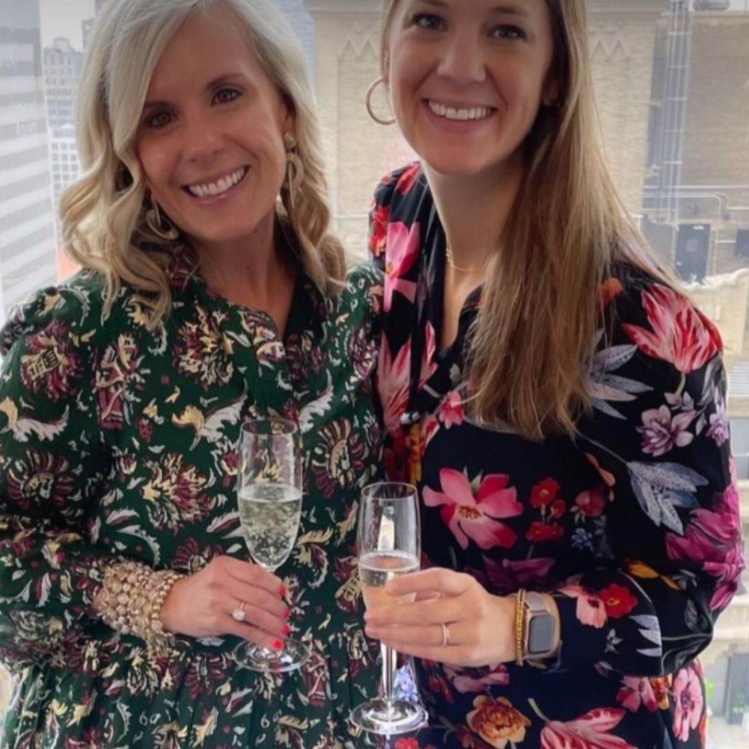 Two women holding drinks and wearing Brett Lauren gemstone bracelets, smiling and posing together in floral outfits.