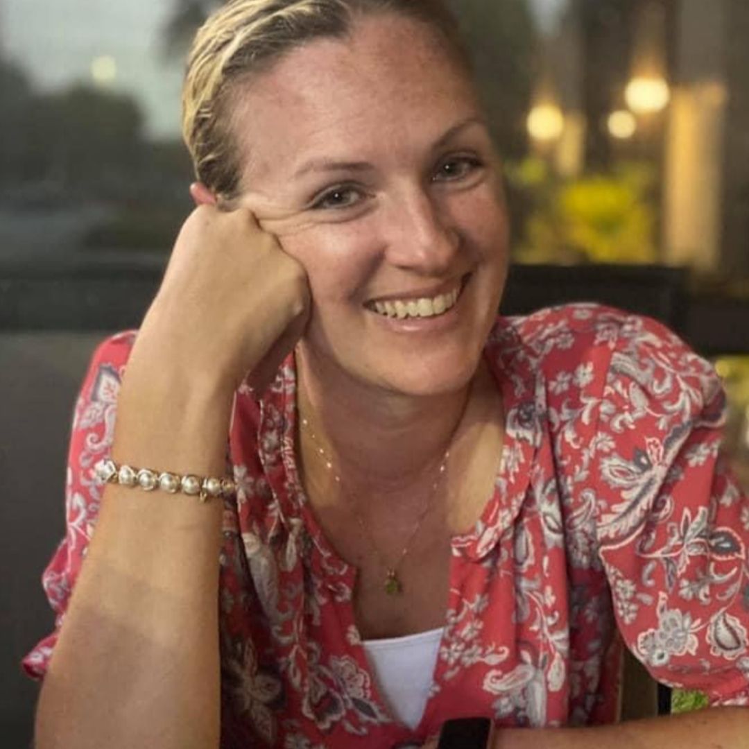 A woman smiling in a casual setting, wearing a Brett Lauren gemstone bracelet, styled with a floral top.