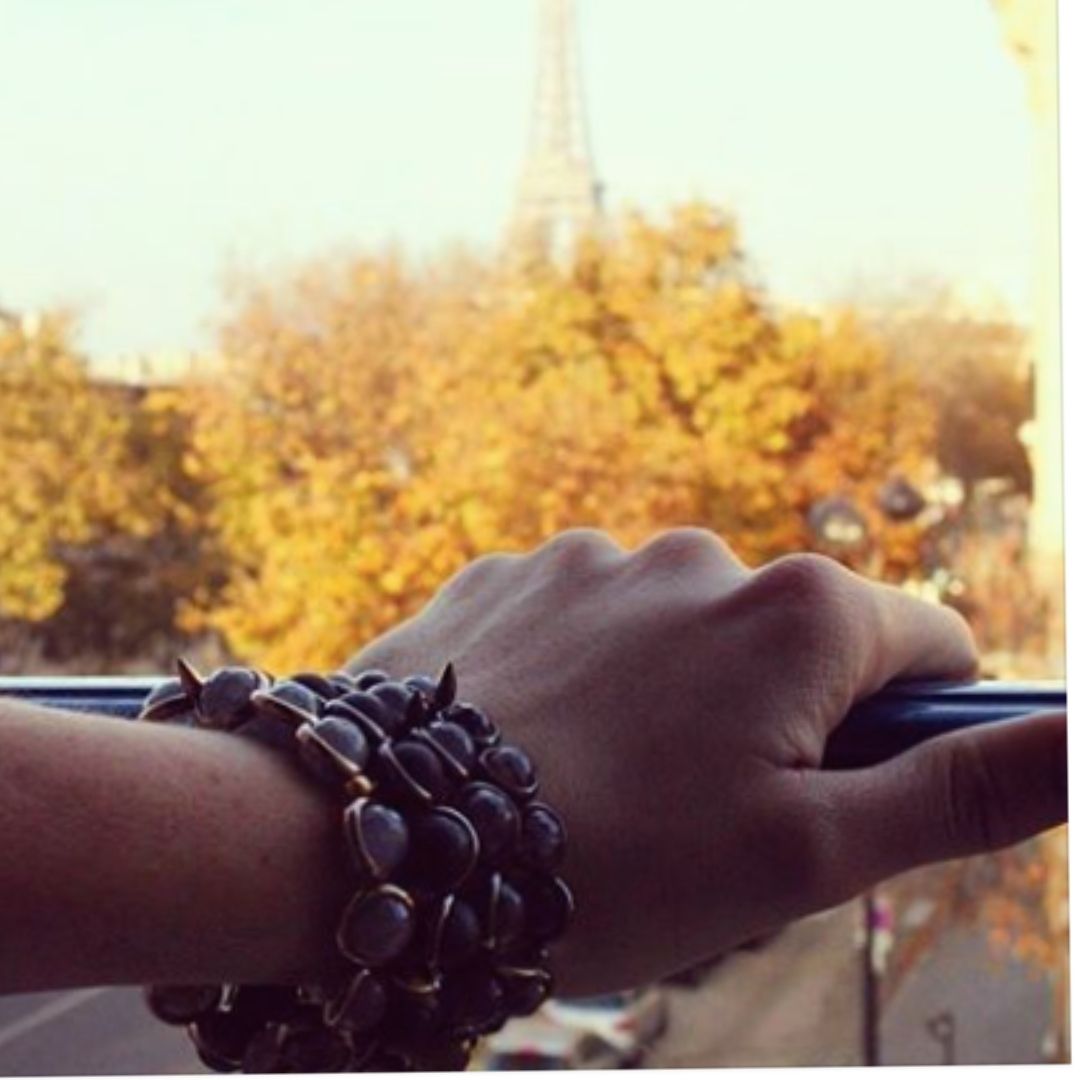 A woman wearing black gemstone bracelets by Brett Lauren while driving, with the Eiffel Tower visible in the background, capturing a scenic view.