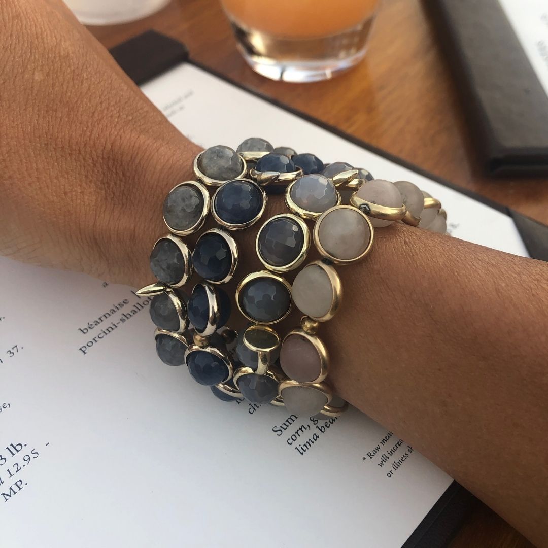 A woman wearing Brett Lauren gemstone bracelets, seated at a table with drinks, showcasing her jewelry in a casual setting.