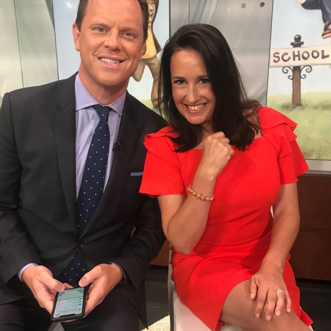 A woman wearing Brett Lauren jewelry sitting next to a man during a TV interview, smiling and posing in a red dress.